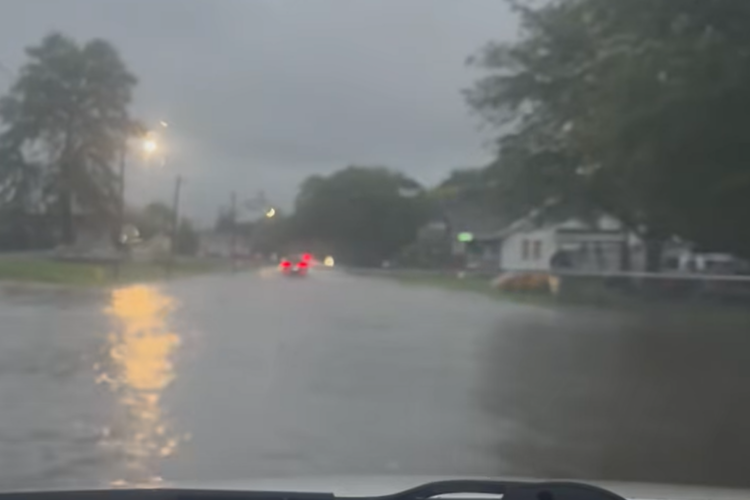 A video taken from the dashboard of a car shows a flooded street with cars driving through standing water below a heavily clouded sky.