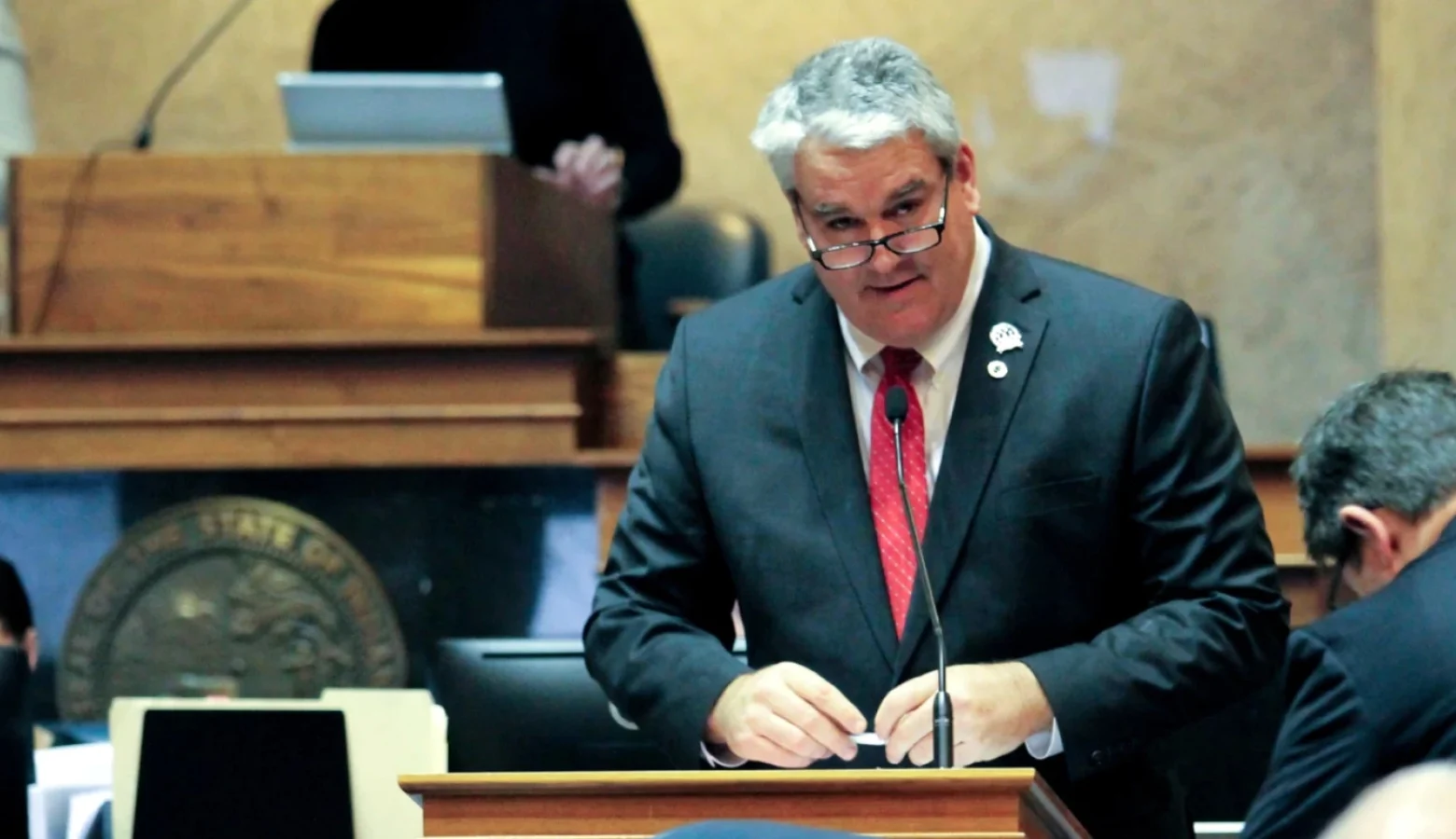 Andy Zay stands at a podium with a mic in a Statehouse chamber. He is wearing a medium suit with a red tie.