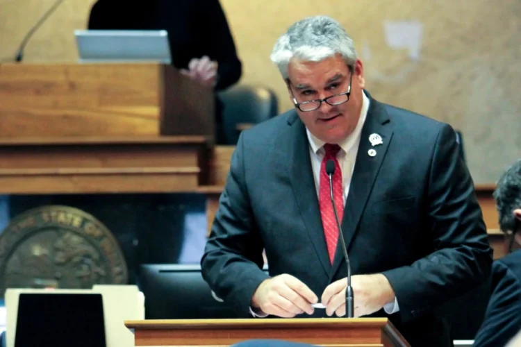 Andy Zay stands at a podium with a mic in a Statehouse chamber. He is wearing a medium suit with a red tie.