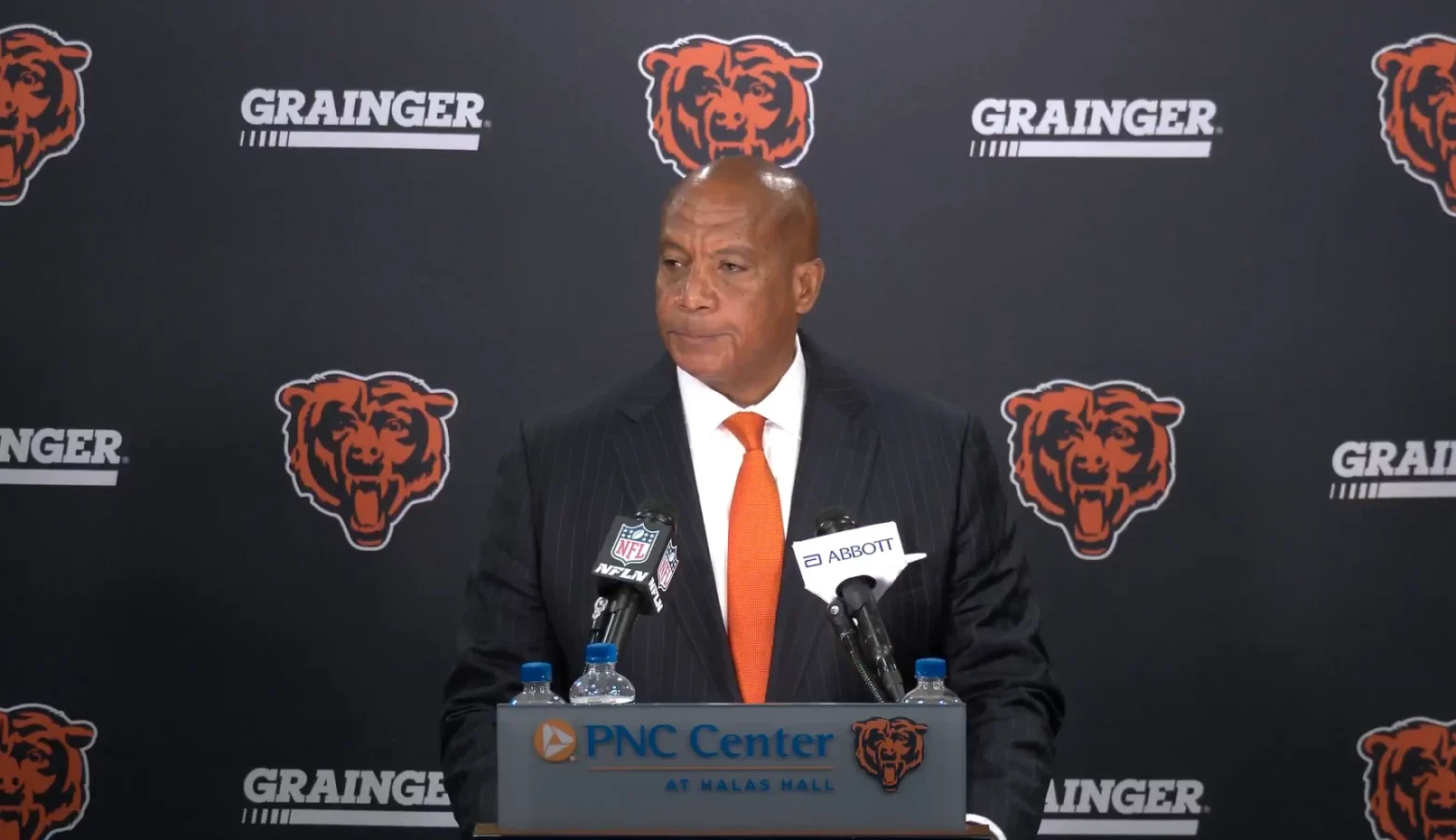 A man in a suit sands at a podium with a microphone. Behind him is a wall featuring the symbol for the Chicago Bears