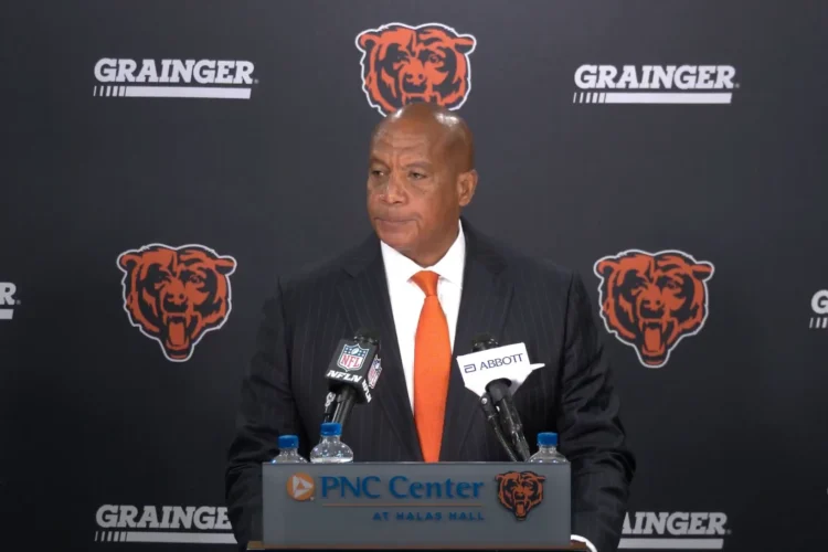 A man in a suit sands at a podium with a microphone. Behind him is a wall featuring the symbol for the Chicago Bears
