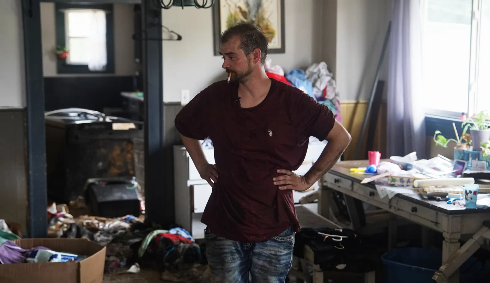 A man stands in a severely messy room. He smokes a cigarette while he observes the area around him