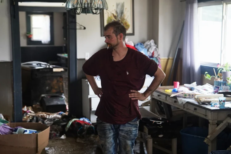 A man stands in a severely messy room. He smokes a cigarette while he observes the area around him
