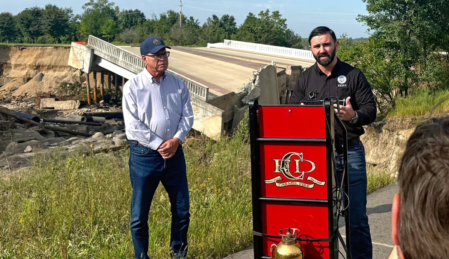 Governor Mike Braun and FEMA administrator Cameron Hamilton stand at a podium set up in a parking lot in front of a bridge destroyed by flood waters.