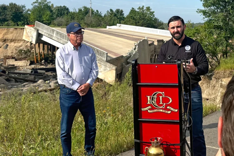 Governor Mike Braun and FEMA administrator Cameron Hamilton stand at a podium set up in a parking lot in front of a bridge destroyed by flood waters.
