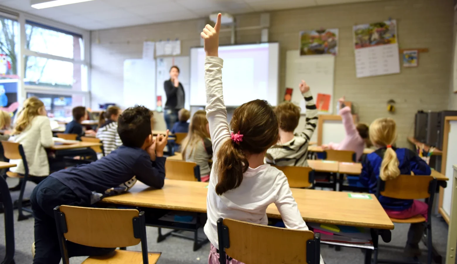 Elementary school students sit at double desks, raising hands to answer a question from a teacher.