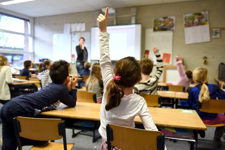 Elementary school students sit at double desks, raising hands to answer a question from a teacher.