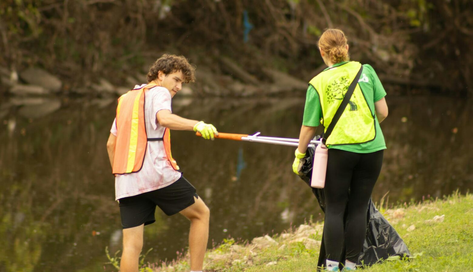 Two volunteers with high-visibility vests use grabber sticks and plastic trash bags to remove debris from the river bank.