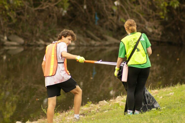 Two volunteers with high-visibility vests use grabber sticks and plastic trash bags to remove debris from the river bank.