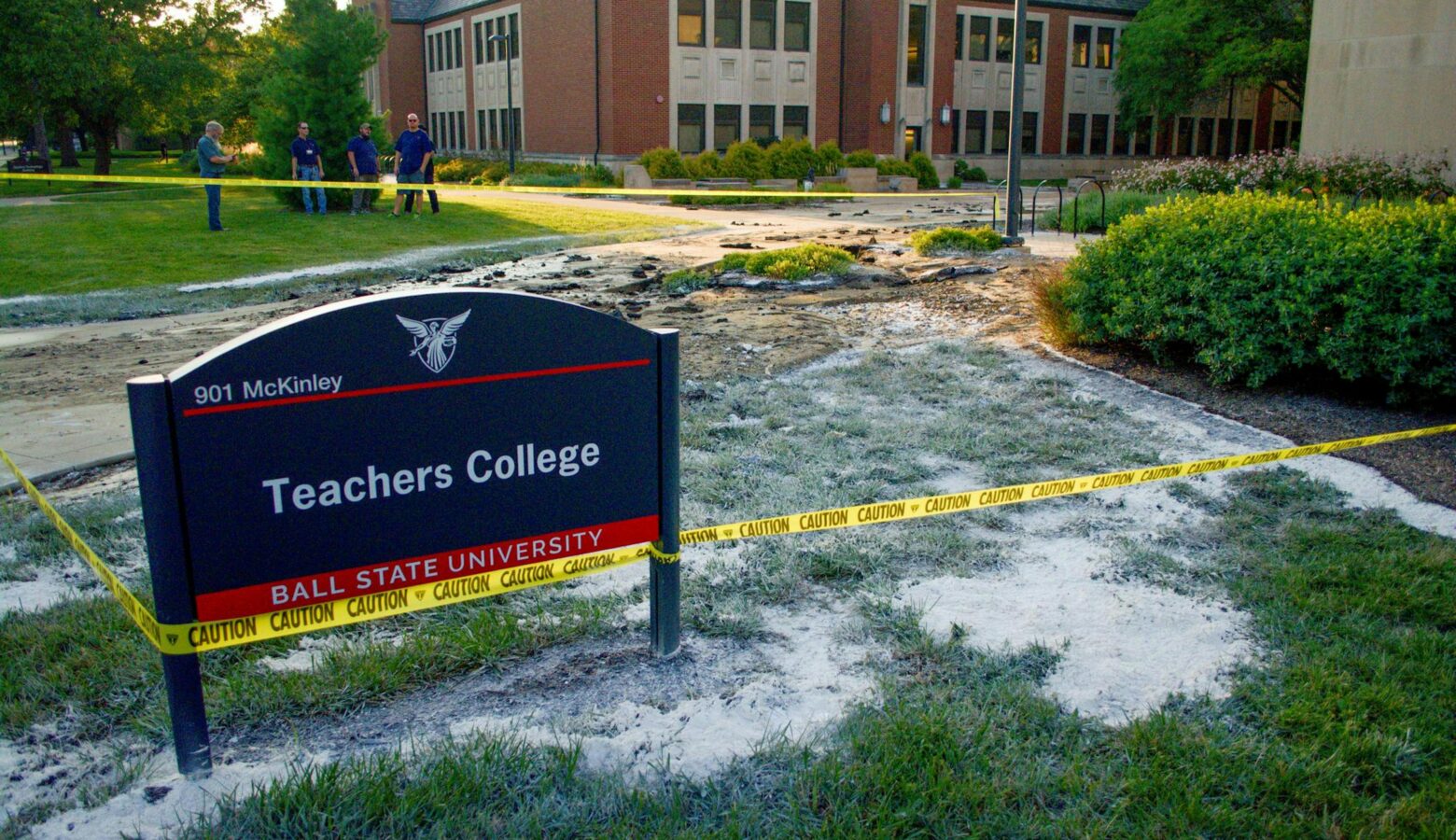 The evening photo shows the Ball State Teachers College sign in the foreground with yellow caution tape around it and extending out and around an area includion grass, shrubs, sidewalks and bicycle racks. The Applied Sciences building is in the background. Four work crew members are discussing the situation outside of the caution tape. Grass and dirt around and near the sign is covered with a white powder.