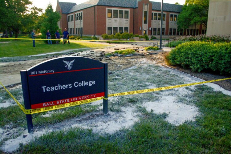 The evening photo shows the Ball State Teachers College sign in the foreground with yellow caution tape around it and extending out and around an area includion grass, shrubs, sidewalks and bicycle racks. The Applied Sciences building is in the background. Four work crew members are discussing the situation outside of the caution tape. Grass and dirt around and near the sign is covered with a white powder.