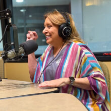 Christina Blanch sits in a radio studio. She is wearing headphones and a multicolored shirt, and is smiling while punching her right fist into the air.