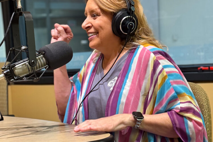 Christina Blanch sits in a radio studio. She is wearing headphones and a multicolored shirt, and is smiling while punching her right fist into the air.