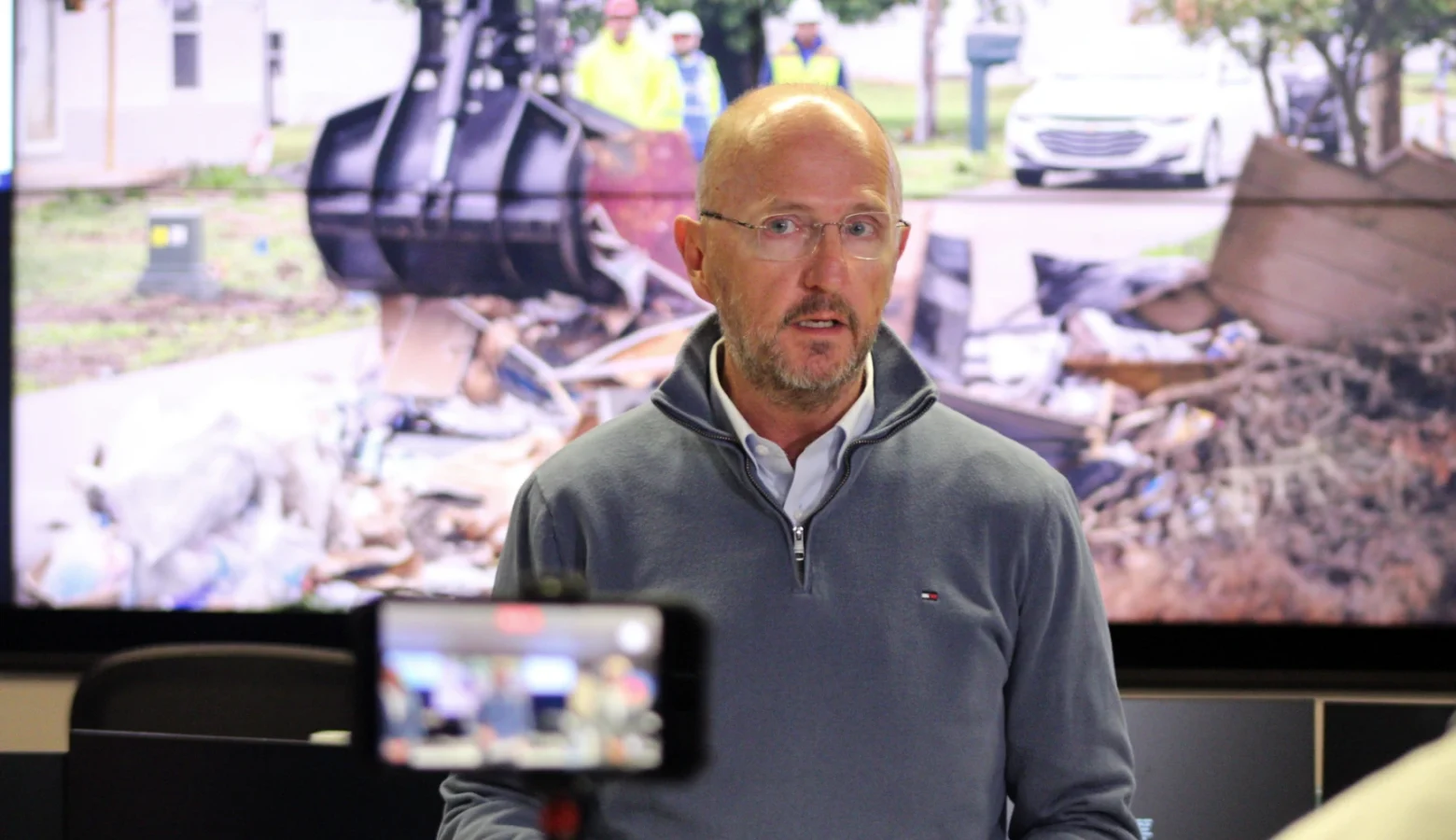 Kevin Guthrie is leading the state’s debris removal task force. He said Indiana’s recovery effort could become the “textbook” for how to manage a natural disaster. Guthrie is dressed in a gray sweatshirt standing in front of a projection screen showing an excavator scooping wreckage.