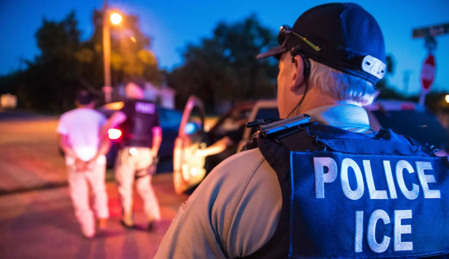 An ICE officer faces away from the camera as he watches another officer arresting someone. Both are walking away from the camera out of focus. The photo was taken on a street either at dusk or dawn.