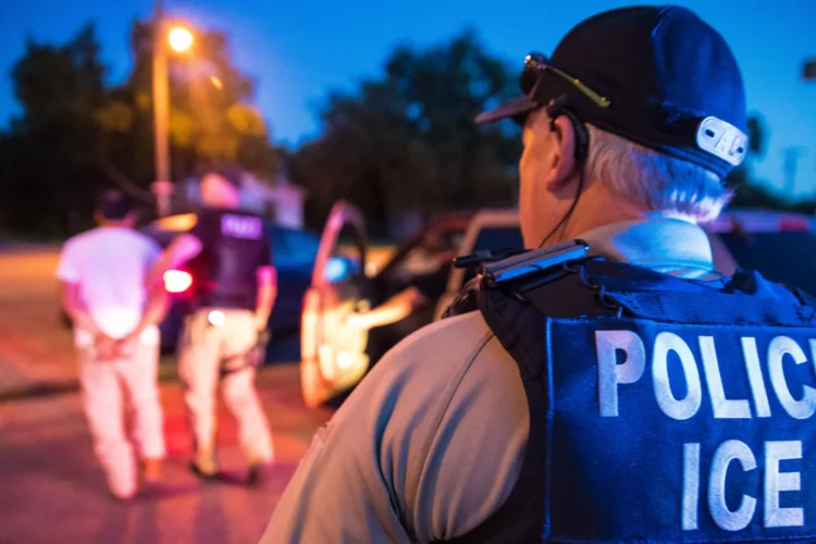 An ICE officer faces away from the camera as he watches another officer arresting someone. Both are walking away from the camera out of focus. The photo was taken on a street either at dusk or dawn.