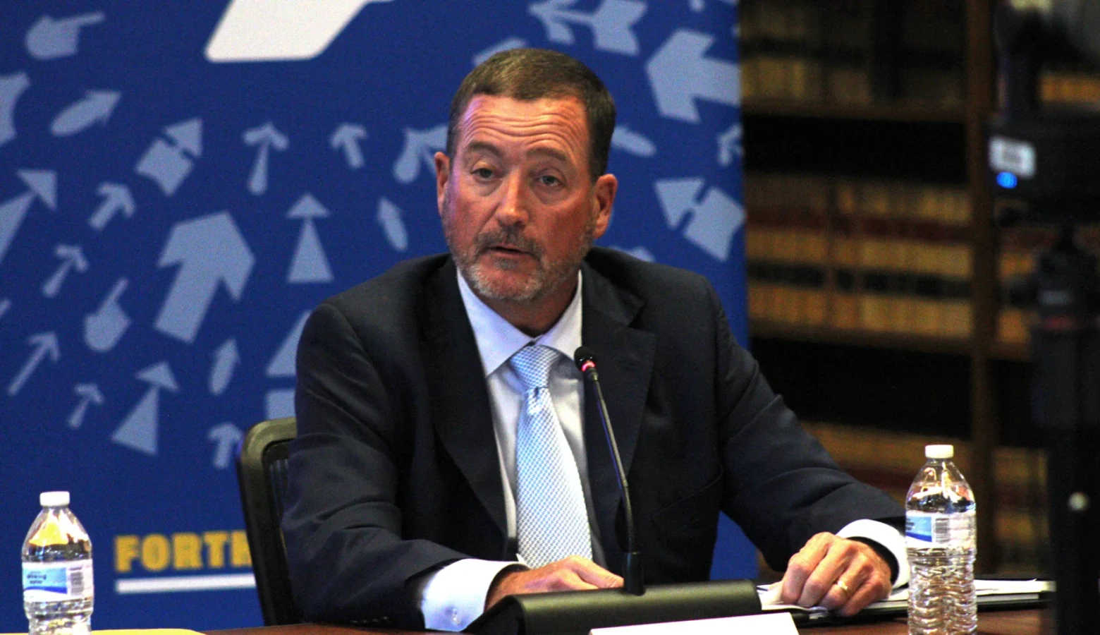 Commerce Secretary Chuck Goodrich discusses the state’s positive economic outlook at Monday’s IEDC meeting at the Indiana State Library September, 28th. He is seated at a table wearing a sport coat and tie with a medium blue background featuring light blue arrows pointing to the upper center of the backdrop.