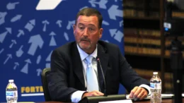 Commerce Secretary Chuck Goodrich discusses the state’s positive economic outlook at Monday’s IEDC meeting at the Indiana State Library September, 28th. He is seated at a table wearing a sport coat and tie with a medium blue background featuring light blue arrows pointing to the upper center of the backdrop.