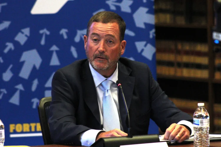 Commerce Secretary Chuck Goodrich discusses the state’s positive economic outlook at Monday’s IEDC meeting at the Indiana State Library September, 28th. He is seated at a table wearing a sport coat and tie with a medium blue background featuring light blue arrows pointing to the upper center of the backdrop.