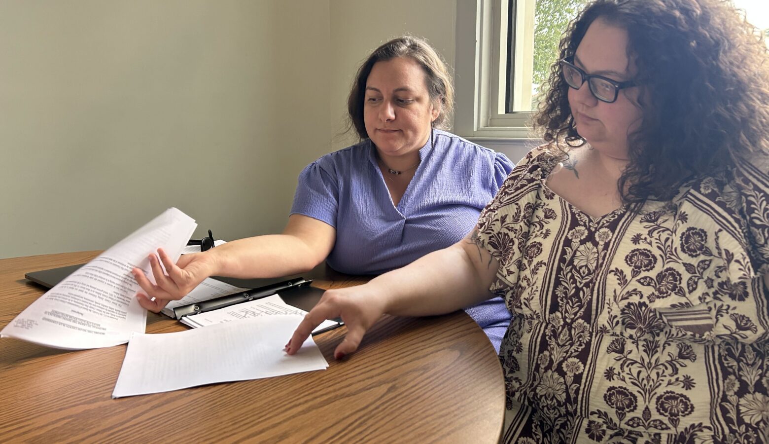 Two women sit at a table, paging through a black binder full of paper.