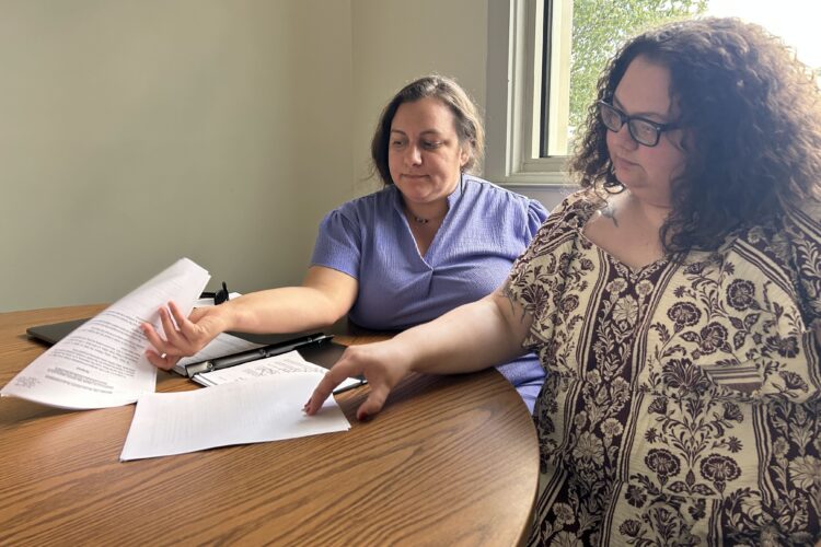 Two women sit at a table, paging through a black binder full of paper.