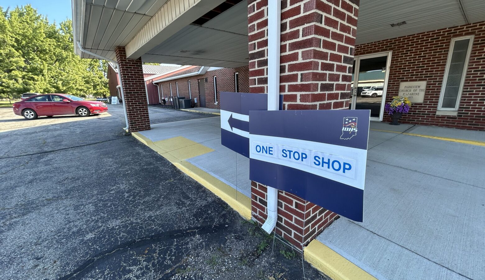 The outside of a brick building. A yard sign is propped up against a brick support. The words "one stop shop" are on it, and it has an arrow pointing left.