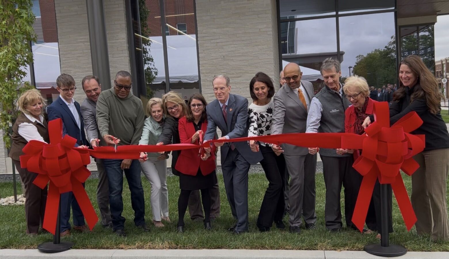 A group of people in professional attire stand behind a bright red ribbon. Several of them hold scissors