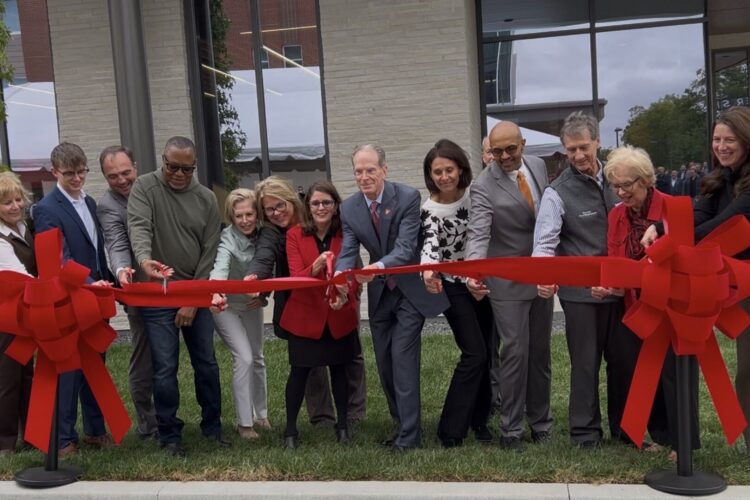 A group of people in professional attire stand behind a bright red ribbon. Several of them hold scissors