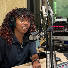KeAnna Mills sits in a radio studio. She has curly hair and is wearing a black polo and headphones on her head. She is talking into a microphone and smiling.