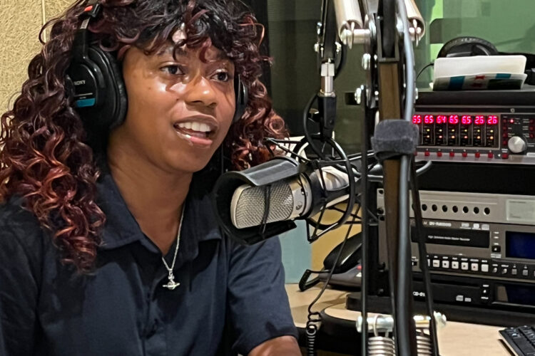 KeAnna Mills sits in a radio studio. She has curly hair and is wearing a black polo and headphones on her head. She is talking into a microphone and smiling.