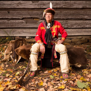 Michael "Mike" Pace sits on furs in traditional Native American dress, including a bright red shirt and a hat with both fur and feathers.