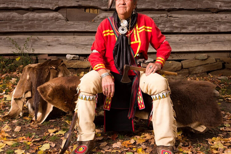 Michael "Mike" Pace sits on furs in traditional Native American dress, including a bright red shirt and a hat with both fur and feathers.