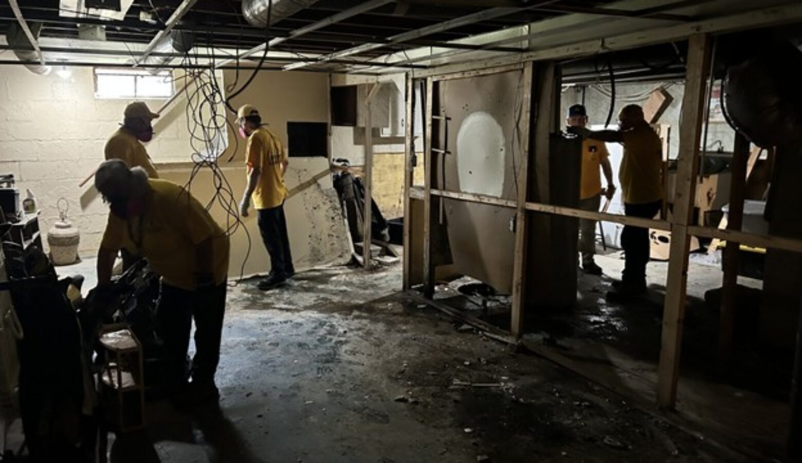 People in yellow t-shirts and hard hats remove paneling from a basement.
