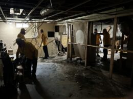 People in yellow t-shirts and hard hats remove paneling from a basement.