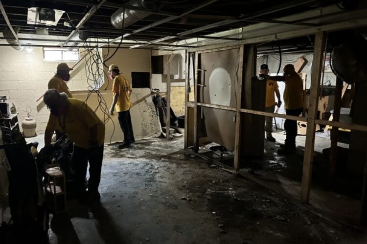 People in yellow t-shirts and hard hats remove paneling from a basement.