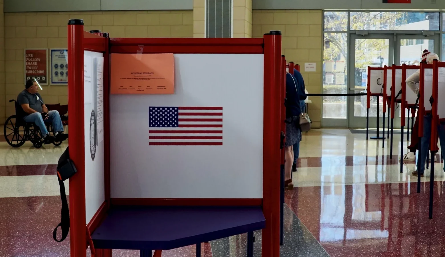 Oct. 5 is the deadline for Indiana residents to register to vote in this year's general election. The photo shows an open and empty voting stand displaying an American flag in a voting precinct. The voting room is brightly lit with sunshine coming through glass doors with bright overhead lights. Voters can be seen in the background occupying other voting booths. A man in a wheelchair is in the distance on the left side of the frame. The room sports a terrazzo floor with burgundy and white areas.