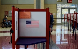 Oct. 5 is the deadline for Indiana residents to register to vote in this year's general election. The photo shows an open and empty voting stand displaying an American flag in a voting precinct. The voting room is brightly lit with sunshine coming through glass doors with bright overhead lights. Voters can be seen in the background occupying other voting booths. A man in a wheelchair is in the distance on the left side of the frame. The room sports a terrazzo floor with burgundy and white areas.
