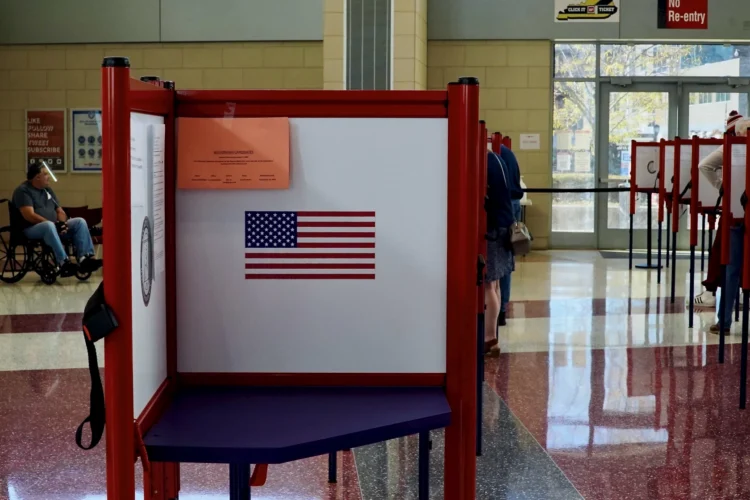 Oct. 5 is the deadline for Indiana residents to register to vote in this year's general election. The photo shows an open and empty voting stand displaying an American flag in a voting precinct. The voting room is brightly lit with sunshine coming through glass doors with bright overhead lights. Voters can be seen in the background occupying other voting booths. A man in a wheelchair is in the distance on the left side of the frame. The room sports a terrazzo floor with burgundy and white areas.