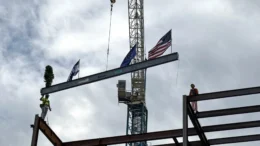 A steel beam is lifted into place by a large crane. The beam has attached flags of the U.S. and Indiana.