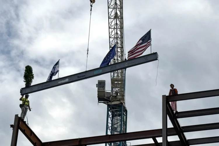 A steel beam is lifted into place by a large crane. The beam has attached flags of the U.S. and Indiana.