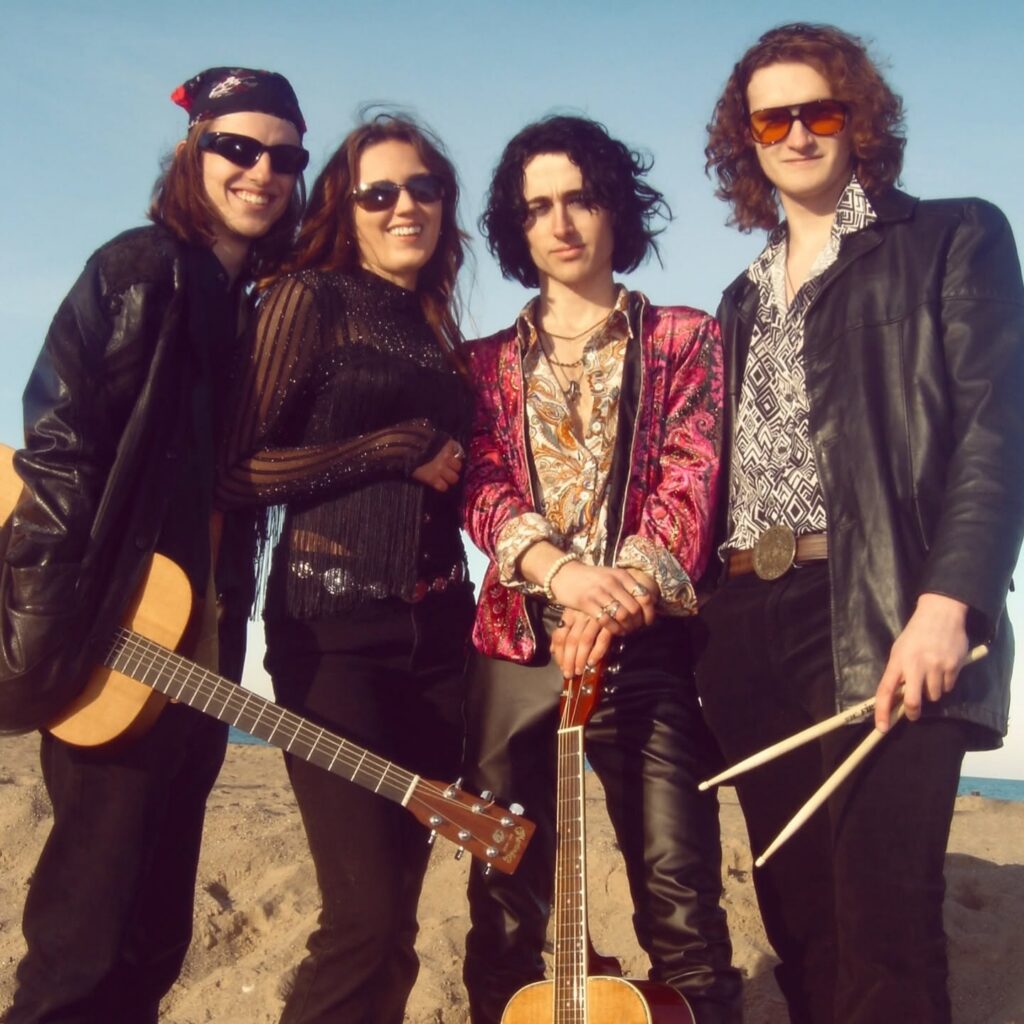 Four band members stand together on a beach, holding musical instruments and facing the camera.