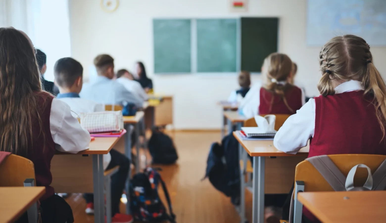 Students, seen from behind, wear school uniforms as they sit in desks in a classroom.