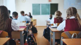 Students, seen from behind, wear school uniforms as they sit in desks in a classroom.