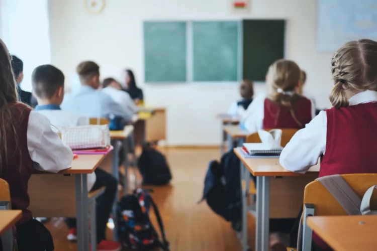Students, seen from behind, wear school uniforms as they sit in desks in a classroom.