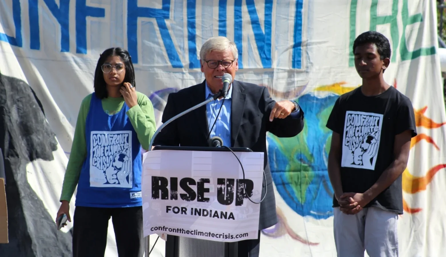 Sen. Ron Alting (R-Lafayette) at a student activist event in 2021, supporting legislative action around climate change. Our photo shows Alting speaking outdoors behind a podium with a "RISE UP FOR INDIANA" banner affixed to it. He is flanked by two students wearing shirts depicting the climate crisis.