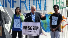 Sen. Ron Alting (R-Lafayette) at a student activist event in 2021, supporting legislative action around climate change. Our photo shows Alting speaking outdoors behind a podium with a "RISE UP FOR INDIANA" banner affixed to it. He is flanked by two students wearing shirts depicting the climate crisis.