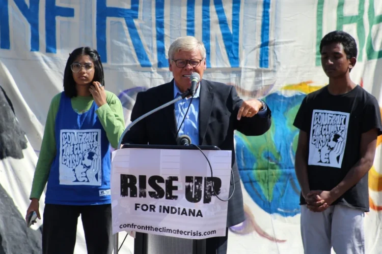 Sen. Ron Alting (R-Lafayette) at a student activist event in 2021, supporting legislative action around climate change. Our photo shows Alting speaking outdoors behind a podium with a "RISE UP FOR INDIANA" banner affixed to it. He is flanked by two students wearing shirts depicting the climate crisis.