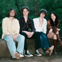 The members of Free Joel; four young people (three men, one woman) sit on a concrete bench with dense greenery behind them. They are wearing casual clothes and neutral expressions.