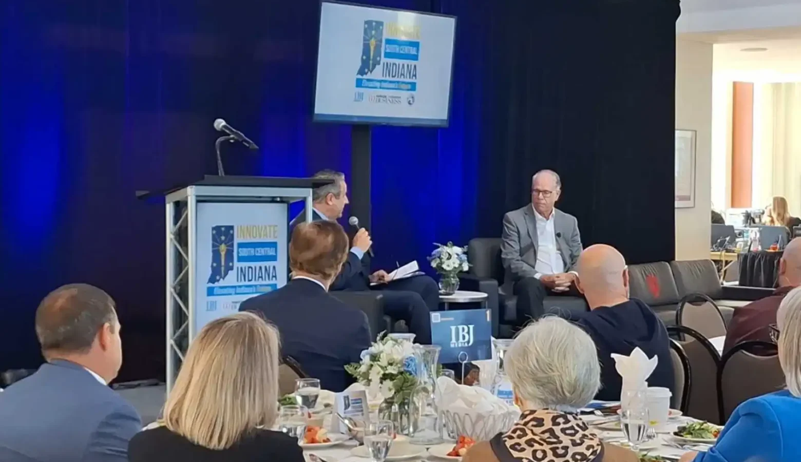 The photo shows a gathering in a conference room with a blue backdrop with a sign displaying the Innovate Indiana logo. Attendees are seated at a roundtable in the foreground while Gov. Braun and a Master of Ceremonies are seated in front of the attendees with a discussion underway.