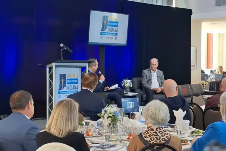 The photo shows a gathering in a conference room with a blue backdrop with a sign displaying the Innovate Indiana logo. Attendees are seated at a roundtable in the foreground while Gov. Braun and a Master of Ceremonies are seated in front of the attendees with a discussion underway.